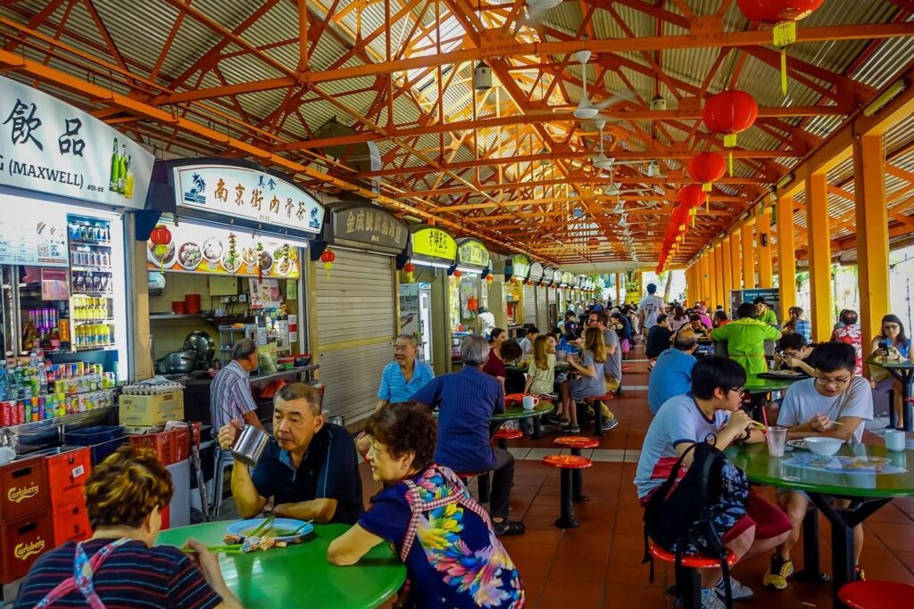 This vibrant image captures a bustling Singaporean hawker center, where people are seated at green circular tables beneath a high, orange-trussed roof adorned with red lanterns. Various food stalls with colorful signage line the left side, serving a diverse crowd of locals and tourists engaged in conversation and dining.