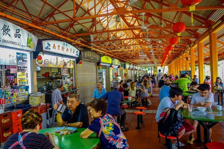 This vibrant image captures a bustling Singaporean hawker center, where people are seated at green circular tables beneath a high, orange-trussed roof adorned with red lanterns. Various food stalls with colorful signage line the left side, serving a diverse crowd of locals and tourists engaged in conversation and dining.