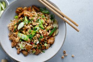 This image features a vibrant bowl of Asian-style noodles topped with savory protein, fresh scallions, and a generous sprinkle of whole peanuts. The dish is presented in a grey ceramic bowl with wooden chopsticks resting on the rim, set against a textured neutral background.