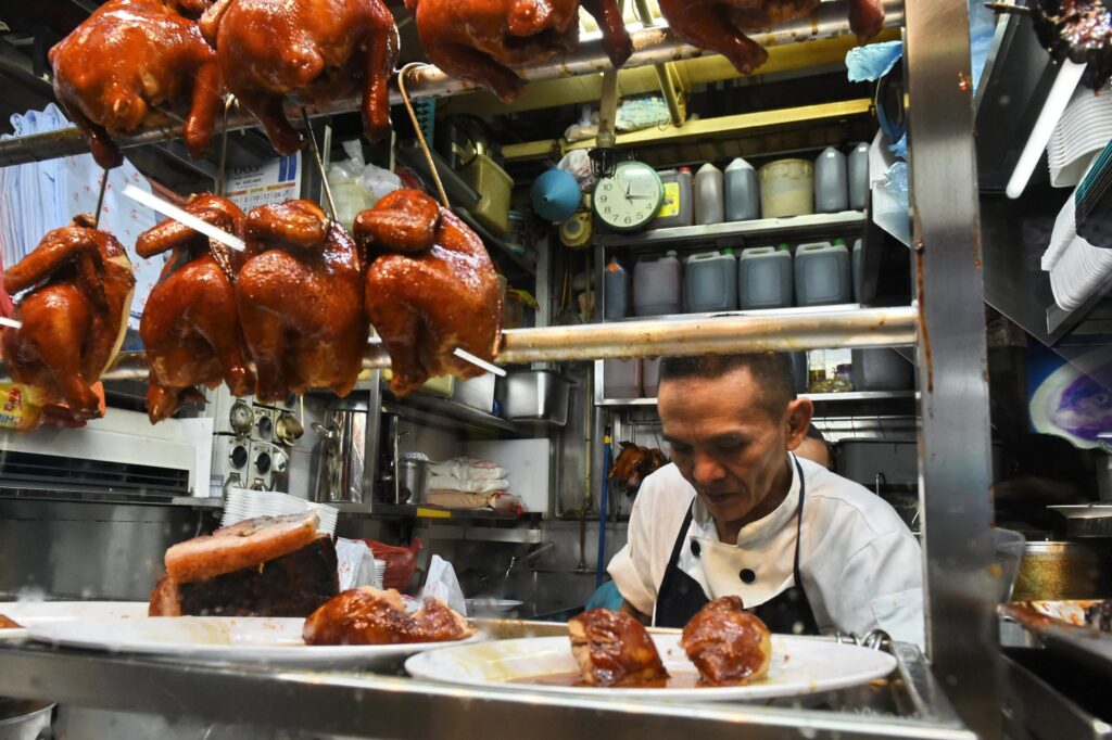 A hawker chef in a white uniform prepares plates of food inside a compact, bustling Singaporean chicken stall. Rows of glistening, roasted whole chickens hang from metal racks above the counter as the centerpiece of the shop.
