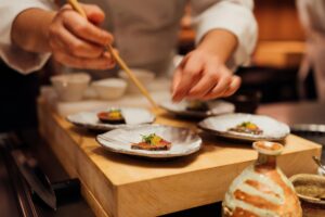 A chef in a white uniform is meticulously plating small appetizers, using long wooden chopsticks to precisely place garnishes on slices of seared fish. The scene is set in a high-end kitchen or sushi bar, featuring elegant ceramic plates arranged on a thick wooden block alongside traditional Japanese pottery.