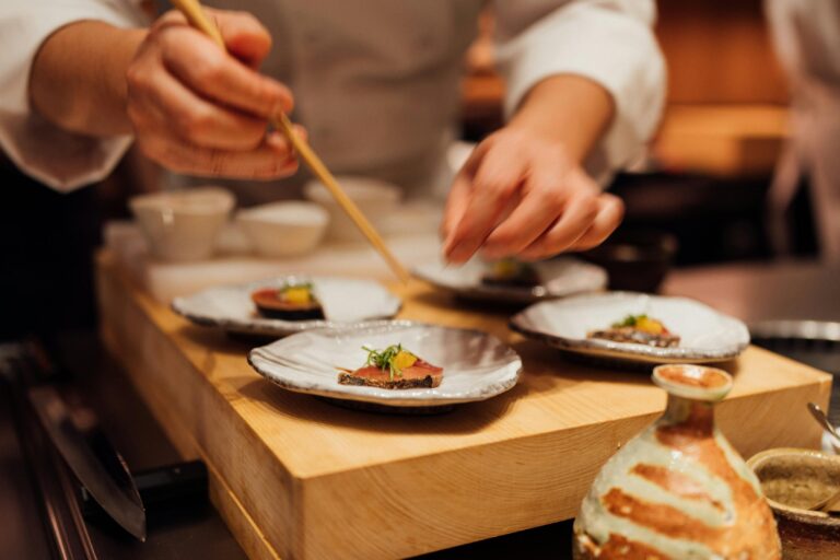 A chef in a white uniform is meticulously plating small appetizers, using long wooden chopsticks to precisely place garnishes on slices of seared fish. The scene is set in a high-end kitchen or sushi bar, featuring elegant ceramic plates arranged on a thick wooden block alongside traditional Japanese pottery.
