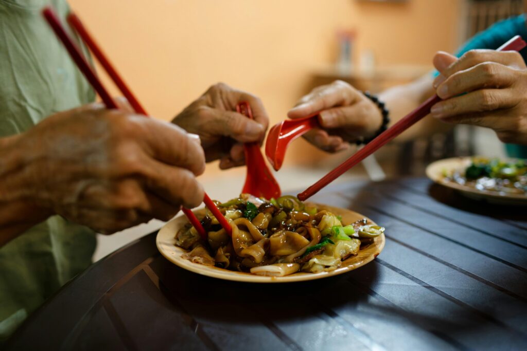 This close-up shot captures multiple people sharing a meal of flat rice noodles, likely char kway teow or hor fun, from a central plate. Using red chopsticks and spoons, the diners reach into the dish, highlighting a communal and social dining experience.
