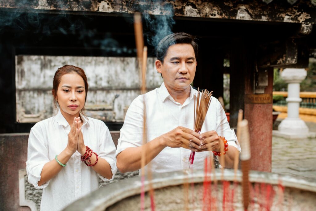 A woman and a man dressed in white shirts are shown in a moment of prayer at a temple, with the man holding a bundle of incense sticks. Smoke from the burning incense rises in the foreground, creating a serene and spiritual atmosphere within the traditional architectural setting.