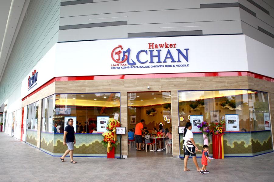 The modern storefront for Hawker Chan is featured with its signature logo and a glass facade showcasing a brightly lit interior. Pedestrians walk past the restaurant's entrance, which is decorated with celebratory flower stands.