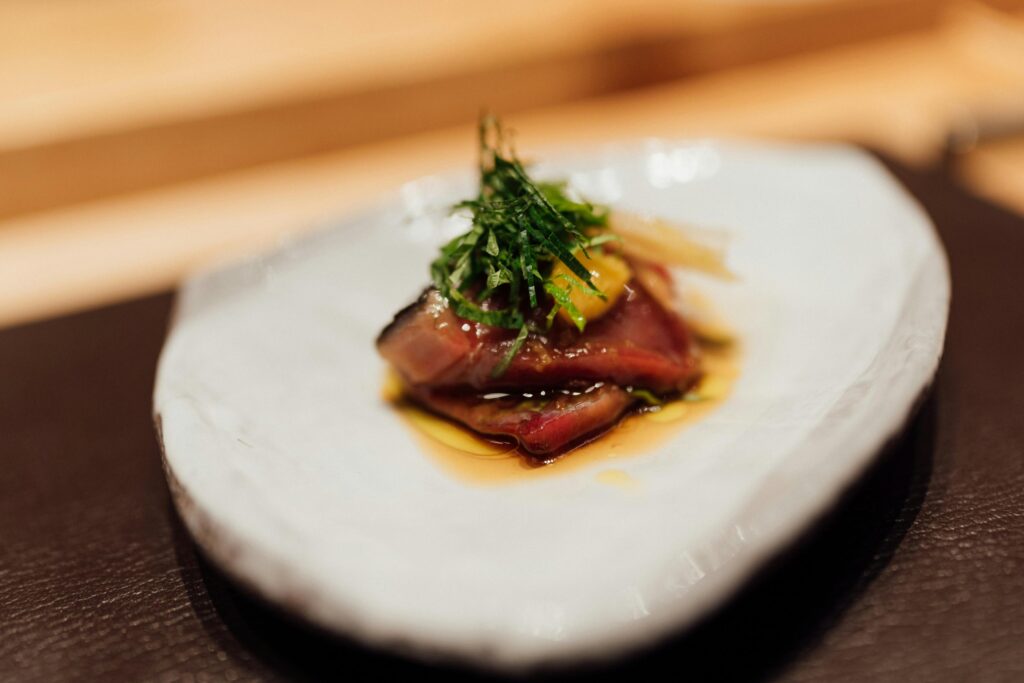 This close-up shot features a delicate serving of seared fish, likely tuna or bonito, resting in a shallow pool of savory sauce on a rustic white ceramic plate. The dish is topped with a bundle of finely shredded green herbs and a touch of citrus zest, emphasizing a minimalist and refined culinary presentation.
