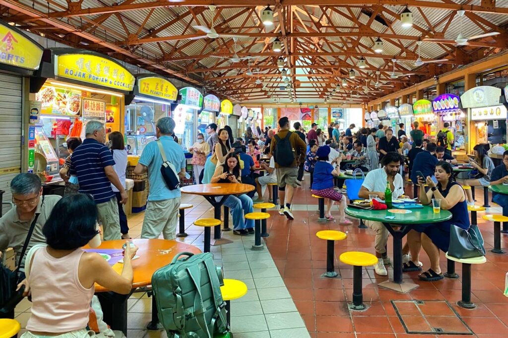 This image shows a bustling, open-air hawker center in Singapore, filled with people dining at colorful round tables beneath a high industrial ceiling. The background features various food stalls with bright yellow signage, advertising local dishes like rojak, popiah, and peanut soup.