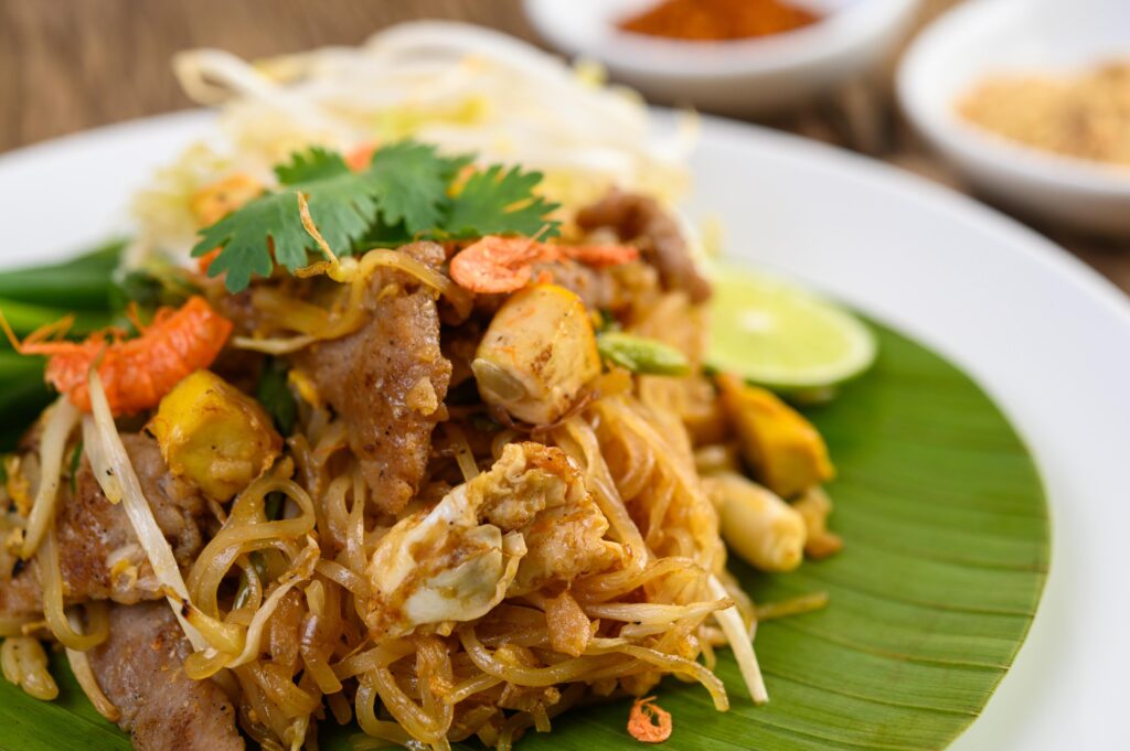 This plate features a vibrant serving of Pad Thai, garnished with fresh cilantro, bean sprouts, and small dried shrimp on a green banana leaf. The noodles are accompanied by a slice of lime and served on a white plate atop a rustic wooden table.