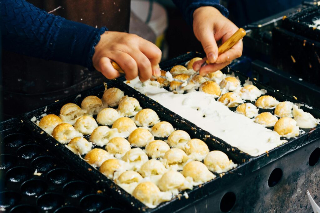 A cook uses wooden-handled picks to skillfully flip golden-brown takoyaki balls within a specialized hemispherical griddle. The pan is filled with a pale batter that is being transformed into spherical street food snacks.