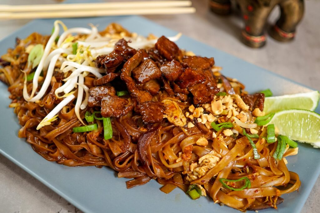 This plate features a mound of dark, stir-fried rice noodles topped with glazed pieces of meat and a garnish of bean sprouts and crushed peanuts. The dish is presented on a blue rectangular plate with lime wedges on the side and a pair of chopsticks visible in the blurred background.