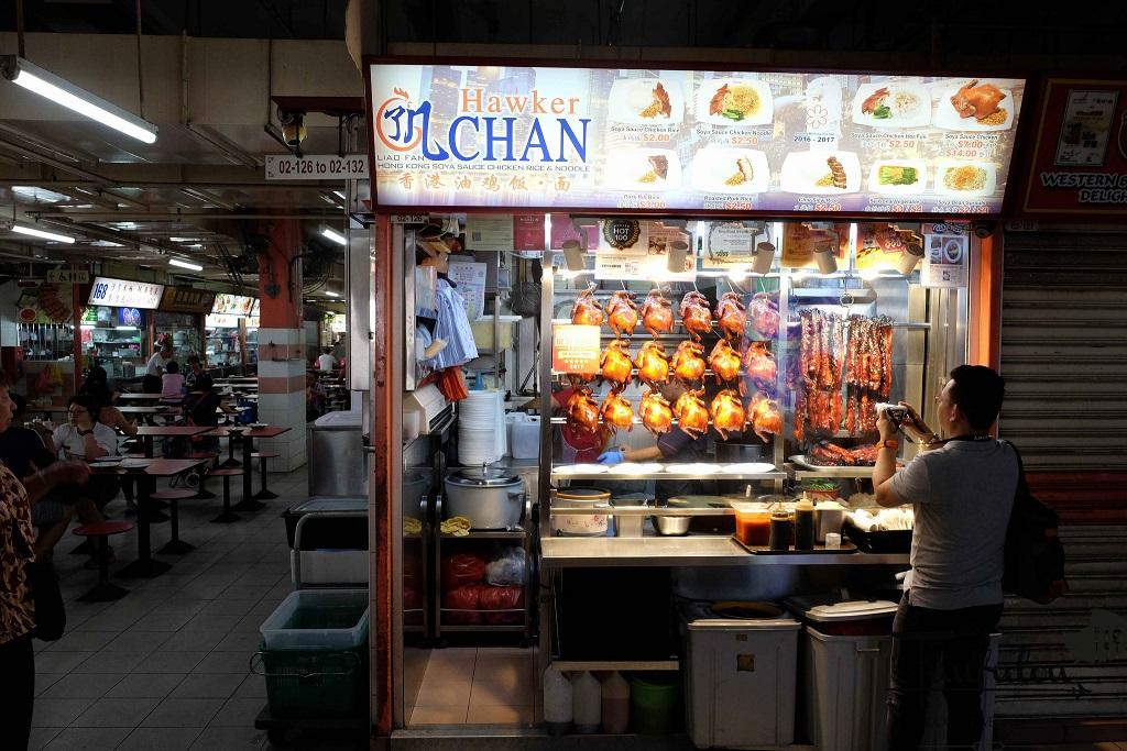A brightly lit Hawker Chan stall stands out in a traditional food center, featuring a large menu board and a customer taking a photograph of the storefront. Behind the glass counter, several rows of signature soy sauce chickens and roasted meats are prominently displayed for diners.