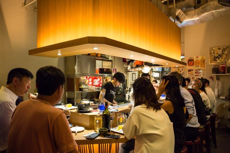This image captures the intimate atmosphere of a Japanese restaurant where diners sit along a wooden counter illuminated by a large, warm overhead light fixture.