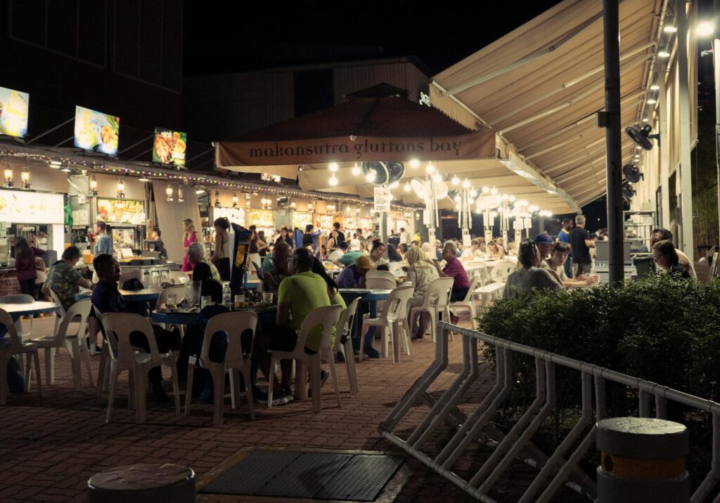 This nighttime scene depicts the Makansutra Gluttons Bay hawker center in Singapore, where diners gather at outdoor tables under warm string lights and large awnings. To the left, several food stalls with bright overhead menus serve a busy crowd of patrons enjoying their meals in the open air.