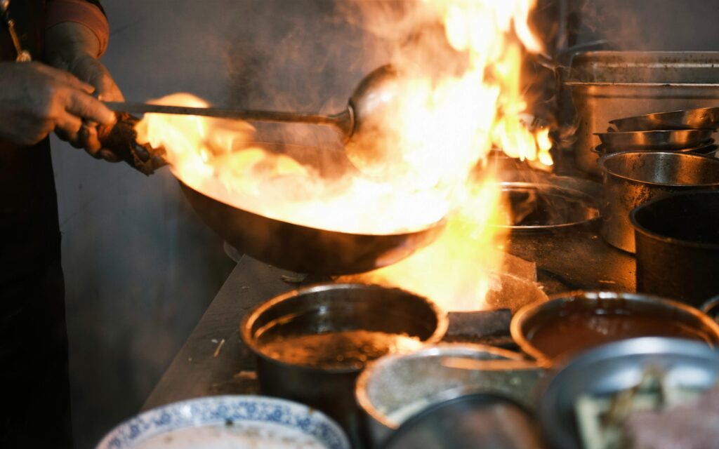 A chef expertly tosses ingredients in a wok over a high-intensity flame, demonstrating the high-heat cooking technique known as "wok hei." The kitchen setting is filled with steam and surrounded by various metal prep containers, capturing the fast-paced energy of a professional culinary environment.