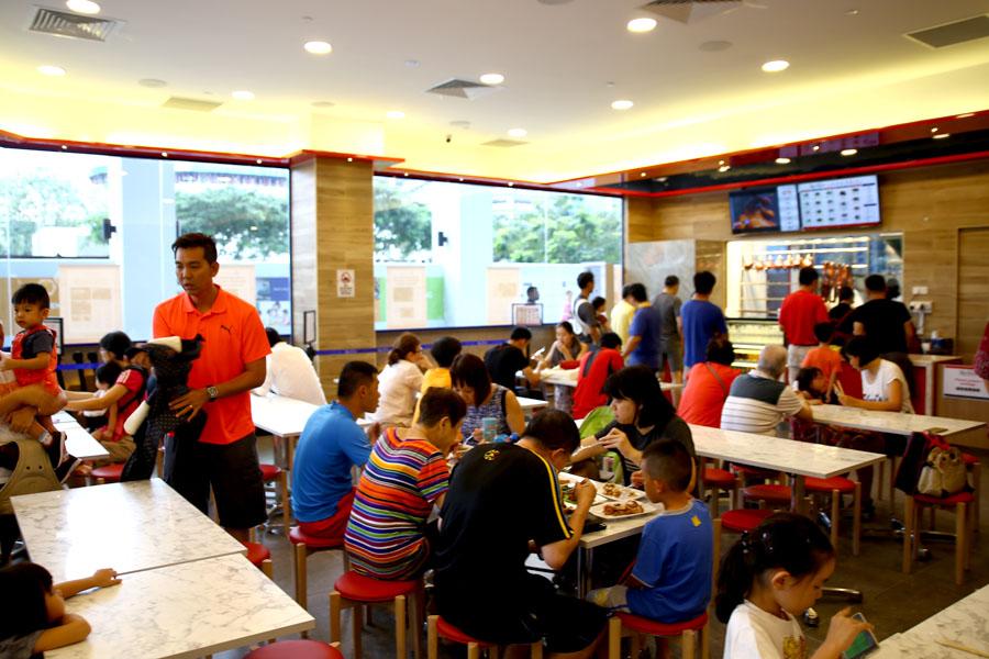 The interior of the Hawker Chan restaurant is filled with diners seated at long tables and red-topped stools. Large windows provide a view of the outside, while a service counter with a digital menu stands at the back of the brightly lit space.