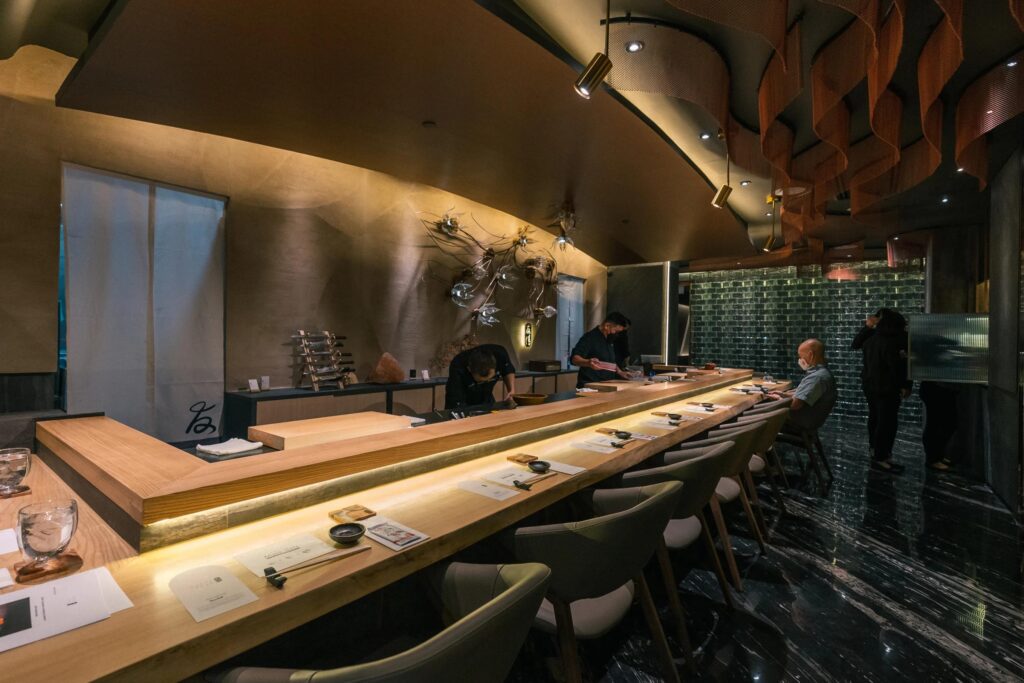 The first image captures a bustling, casual Japanese eatery where diners sit at a wooden counter under a massive, warm light fixture while chefs prepare food in the open kitchen.