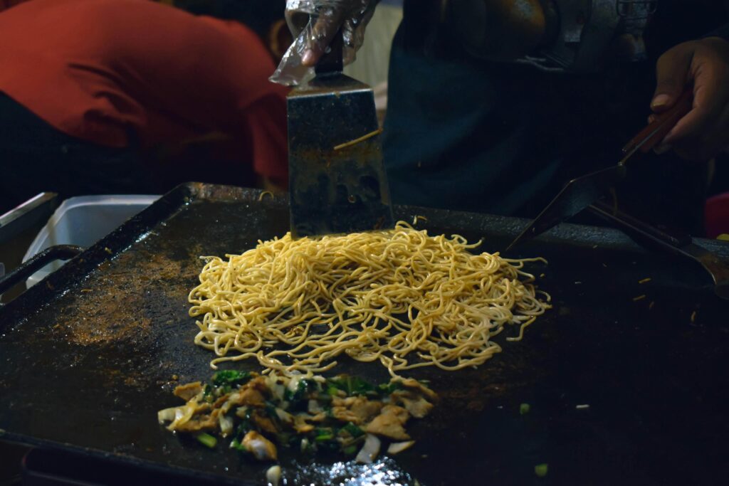 A cook uses large metal spatulas to stir-fry a pile of thin yellow noodles on a seasoned flat-top griddle. In the foreground, a mixture of chopped greens and meat sizzles separately on the dark, oiled cooking surface.
