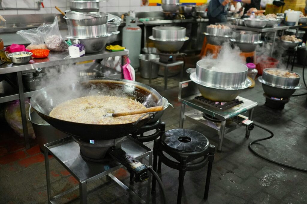 This image showcases a busy commercial kitchen filled with large metal woks and steaming pots arranged on individual gas burners. Various cooking supplies and stainless steel prep tables are scattered throughout the industrial space, highlighting a high-volume food production environment.