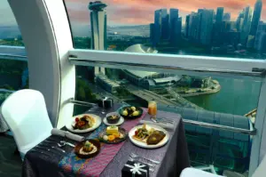 Wide-angle interior shot of a luxury fine dining table inside a glass observation capsule, overlooking Singapore Marina Bay skyline at sunset, with plated gourmet dishes and cityscape in the background.
