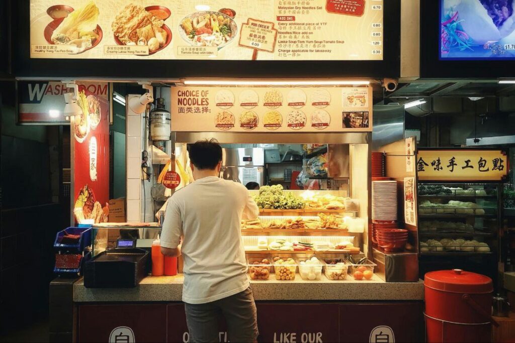 A man in a white t-shirt stands at a brightly lit Yong Tau Foo hawker stall, browsing an array of fresh ingredients and noodle options. Above the counter, colorful menu boards display various soup bases and steamed buns available for order.