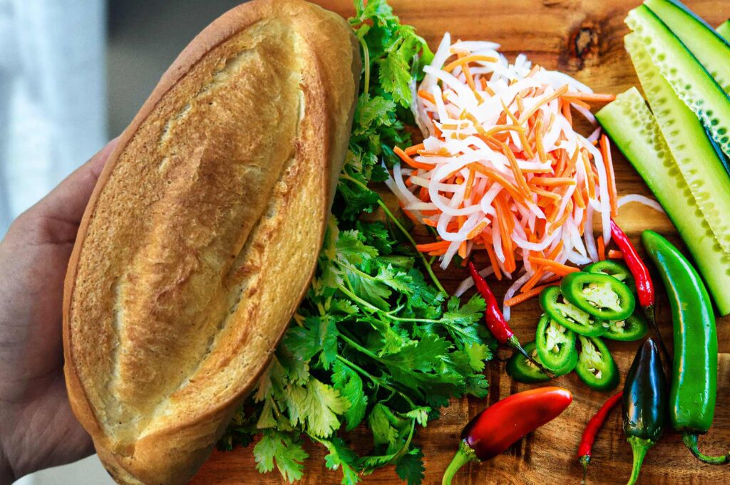 Ingredients for a Vietnamese bánh mì displayed on a wooden board, including a fresh baguette, cilantro, pickled carrots and daikon, sliced cucumbers, jalapeños, and red and green chili peppers.