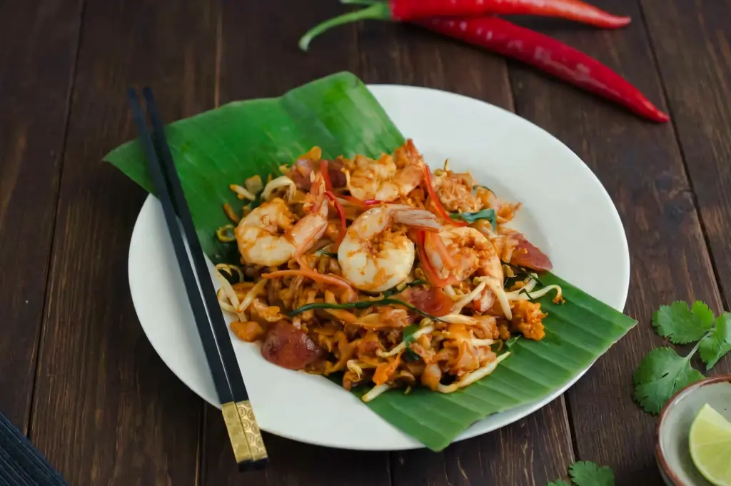 Three‑quarter angle close‑up shot of Penang-style char kway teow served on a banana leaf–lined plate, featuring prawns, noodles, bean sprouts, egg, and chili, styled on a rustic wooden background.