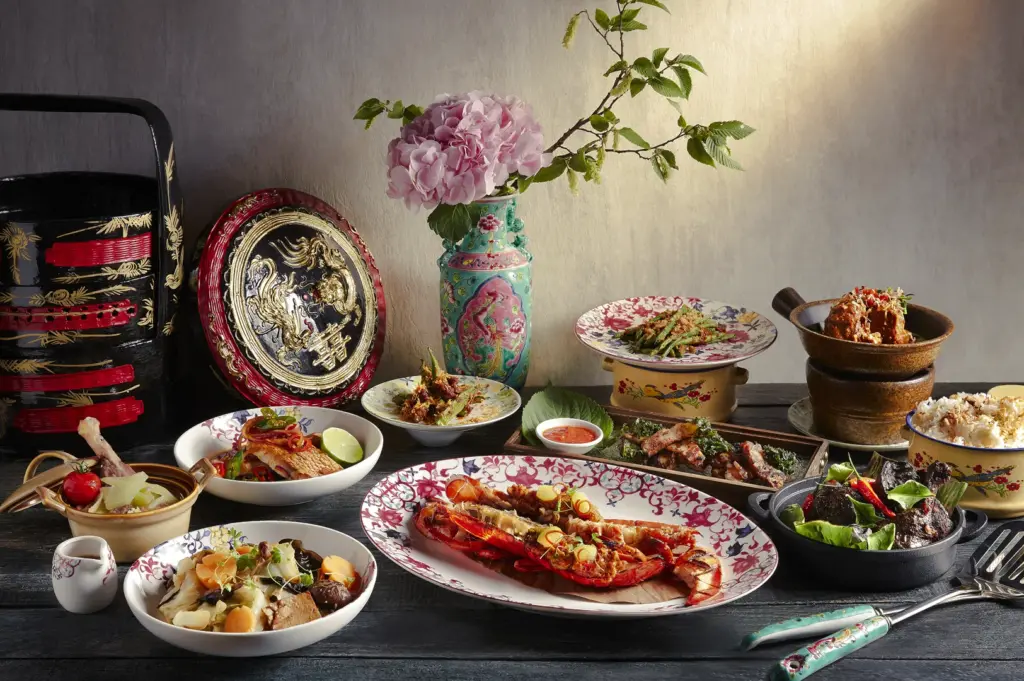 Eye-level shot of an elaborate Asian fine dining spread with lobster, plated entrées, and decorative tableware arranged on a rustic table, styled with floral accents and traditional serving dishes.