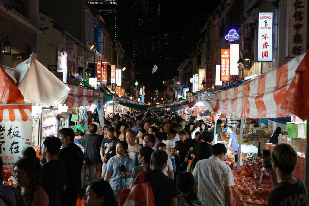 A dense crowd of people navigates a bustling night market lined with vibrant stalls and glowing neon signs. Red and white striped awnings stretch over the vendors, creating a lively atmosphere in this narrow urban corridor.