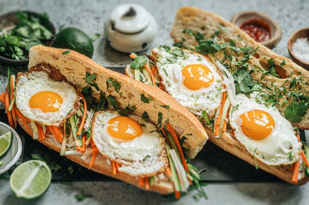 Vietnamese bánh mì sandwiches topped with sunny-side-up eggs, pickled vegetables, cucumbers, fresh herbs, and lime wedges arranged around them on a textured surface.