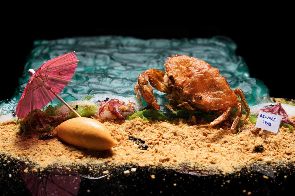 Contemporary gastronomy plate featuring edible crab, foamy surf effect, and beach‑inspired garnishes.