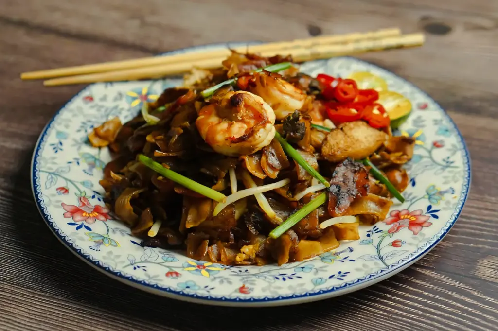 Top‑down close‑up shot of Singapore‑style char kway teow served on a floral ceramic plate, featuring flat rice noodles, prawns, cockles, Chinese sausage, bean sprouts, and chili garnish on a wooden table.