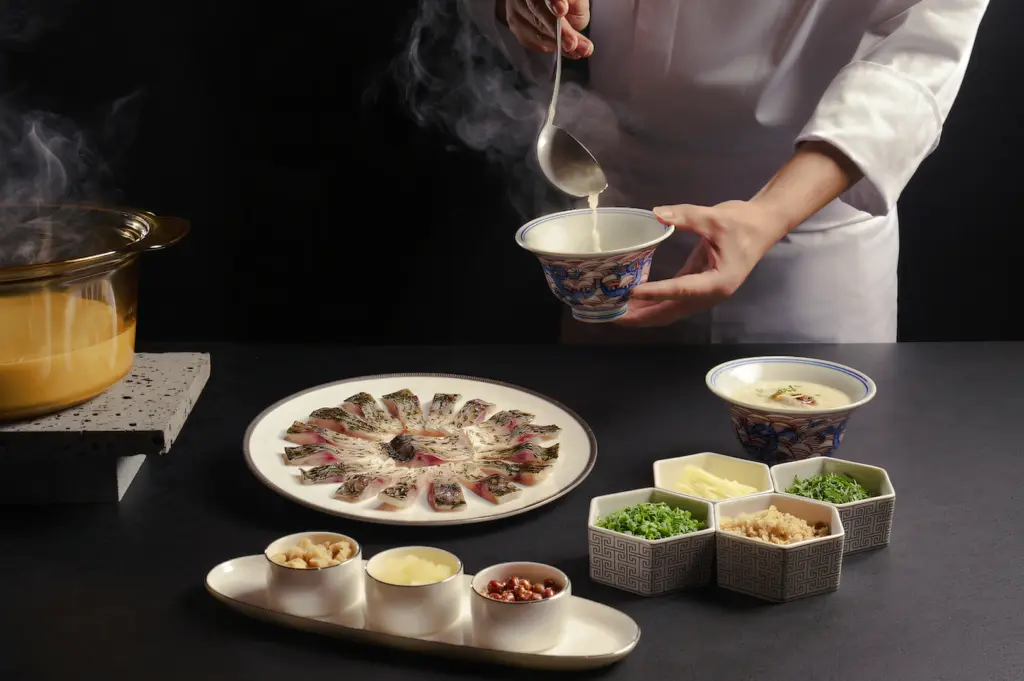 Close-up shot of a chef ladling steaming sauce over an elegantly plated fine dining dish, with garnishes and ingredients arranged on a dark table against a black studio background.