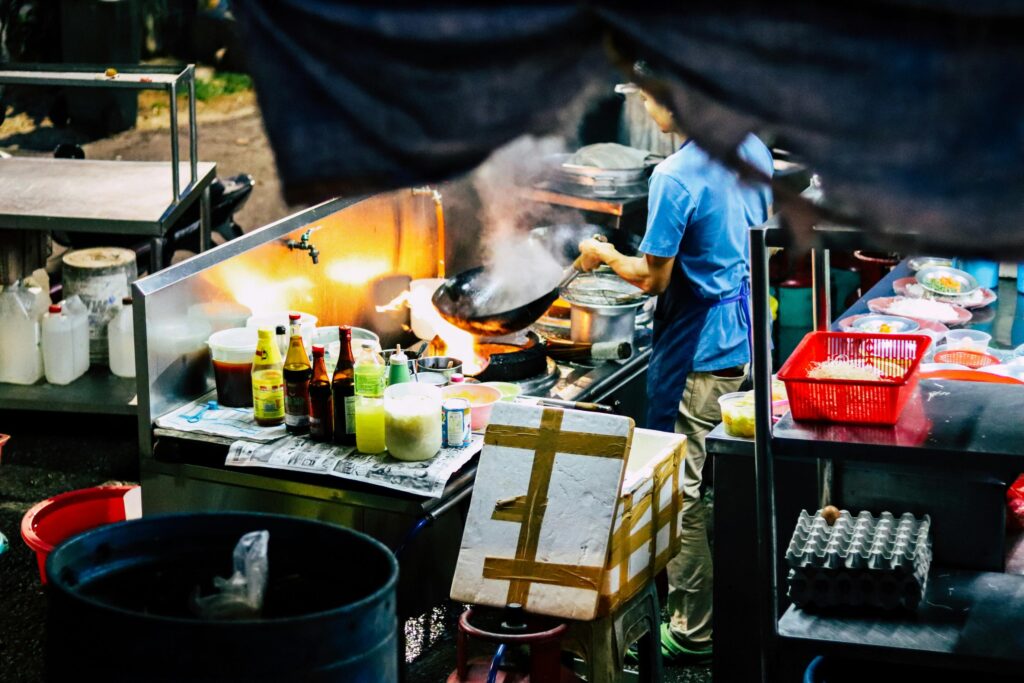 A street food vendor in a blue shirt skillfully tosses ingredients in a wok over a high flame. His compact outdoor kitchen is packed with various sauces, seasonings, and fresh ingredients ready for cooking.