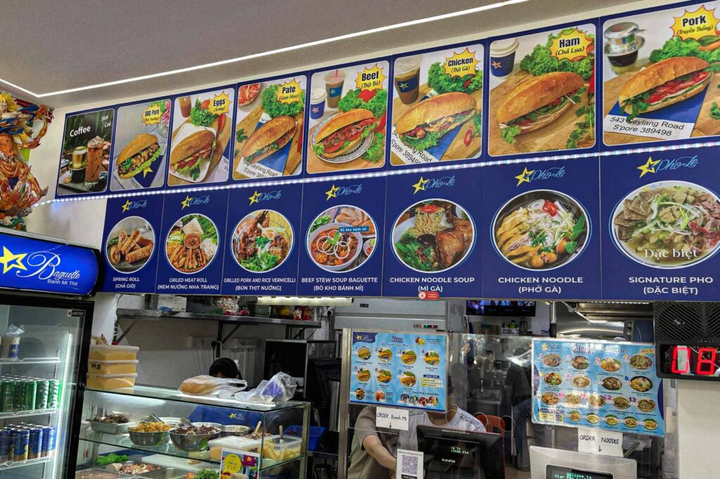 Counter of Star Baguette displaying a menu of bánh mì thịt and other Vietnamese dishes, with large overhead food photos showing different baguette options, noodle soups, and beverages above the service area.