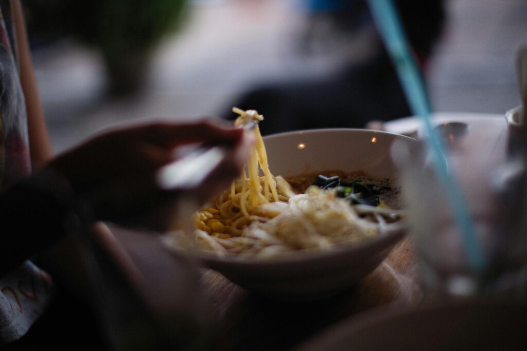 A person uses chopsticks to lift a steaming portion of noodles from a bowl filled with corn and other fresh toppings. The shallow depth of field keeps the focus on the meal, capturing an intimate moment of dining.