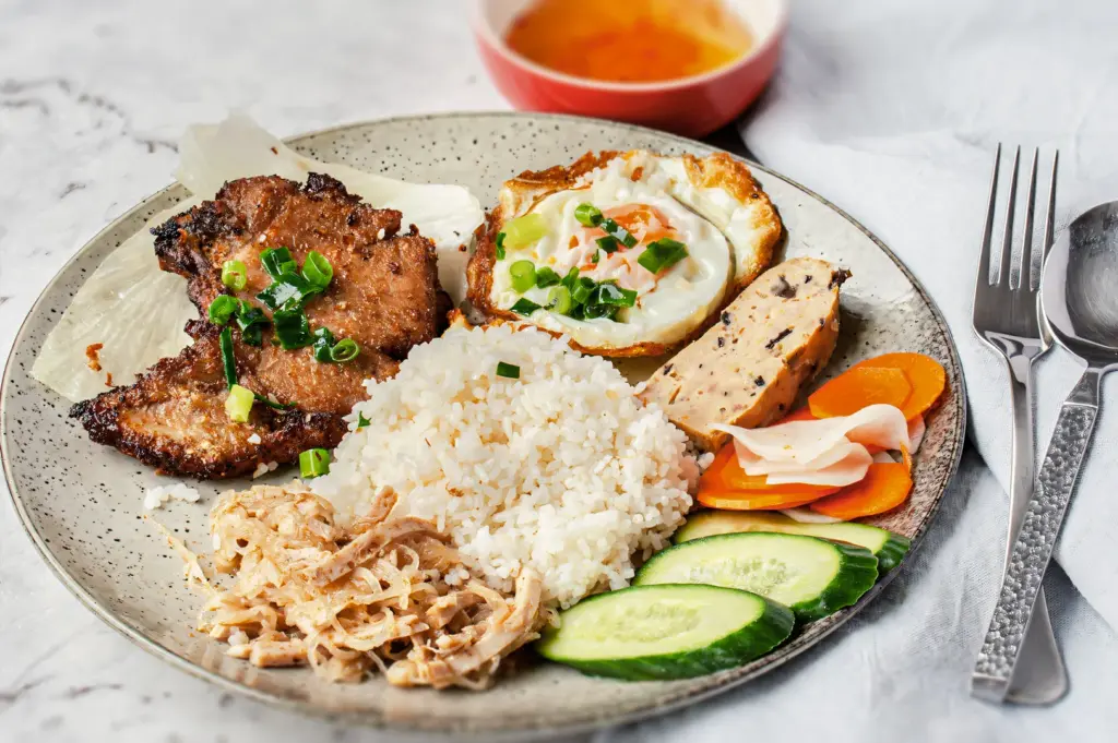 Vietnamese broken rice plate (cơm tấm) with grilled pork, fried egg, pickled vegetables, cucumber slices, and dipping sauce.