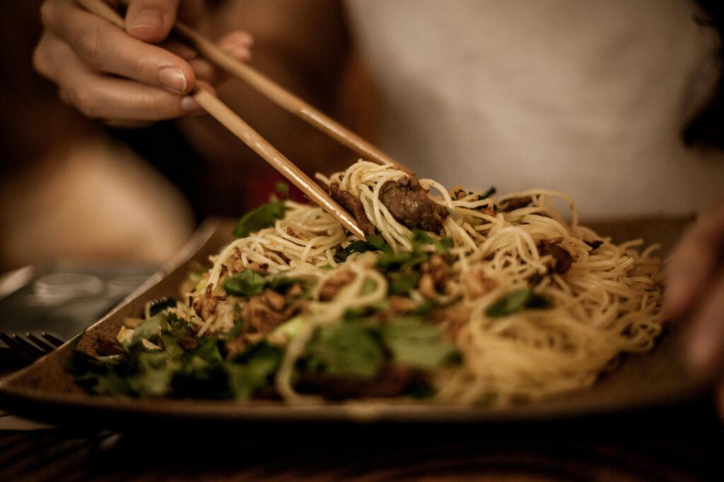 A person uses wooden chopsticks to lift a generous portion of thin noodles mixed with meat and fresh herbs from a dark plate. The warm, soft lighting emphasizes the texture of the dish while keeping the background in a gentle blur.