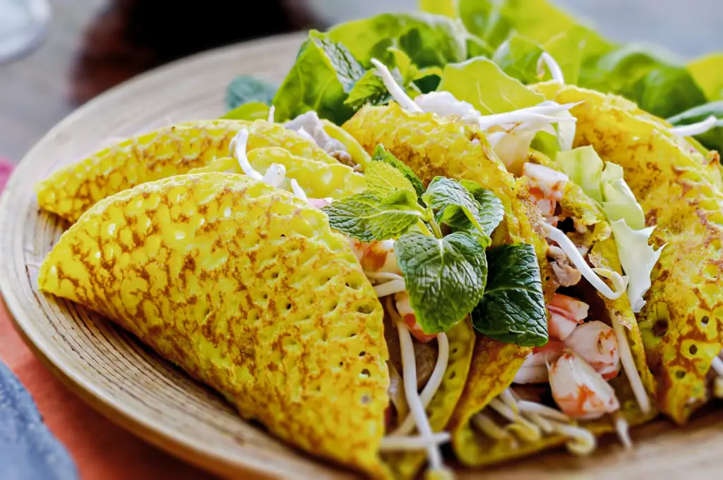 Vietnamese turmeric pancakes (bánh xèo) filled with shrimp, bean sprouts, and fresh herbs, served on a wooden plate.