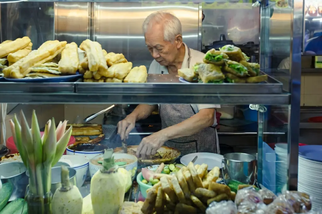 Eye-level medium shot of an elderly Asian hawker preparing traditional street food behind a glass display, stacks of freshly made snacks in the foreground inside a busy urban hawker stall.