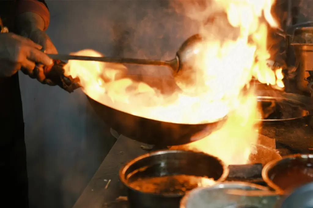 Low-light close-up shot of a chef handling a wok engulfed in flames inside a professional kitchen, demonstrating the intense heat required for authentic wok hei.