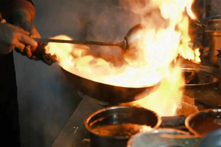 Low-light close-up shot of a chef handling a wok engulfed in flames inside a professional kitchen, demonstrating the intense heat required for authentic wok hei.