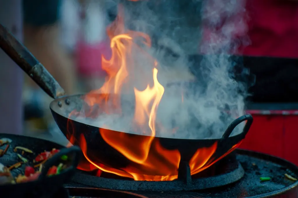 Side‑angle close‑up shot of a metal wok over roaring flames, with thick smoke and fire rising to capture wok hei—the smoky aroma created by high‑heat stir‑frying in Singapore hawker cooking.