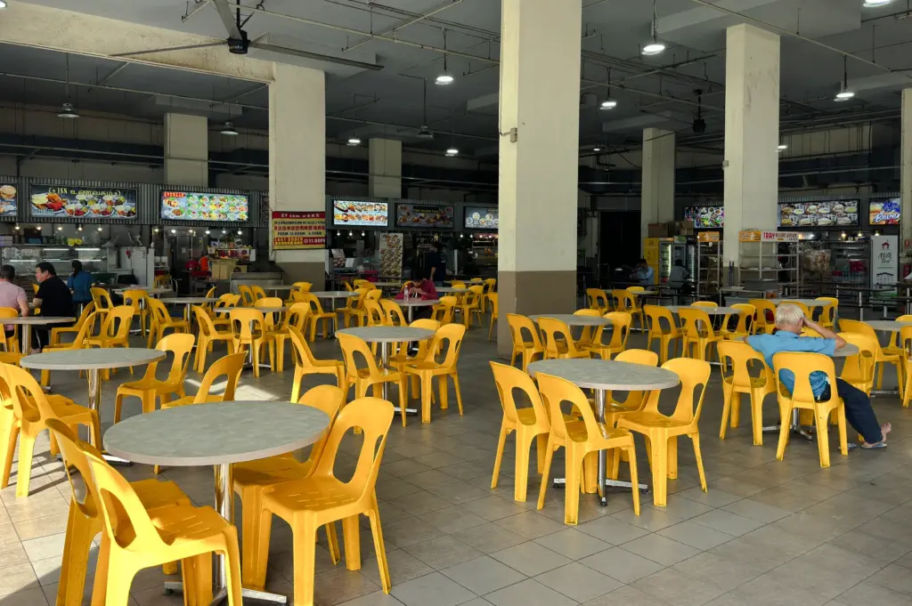 Wide-angle interior shot of a spacious hawker centre with rows of yellow plastic chairs and round tables, showing a quiet dining hall with food stalls in the background.