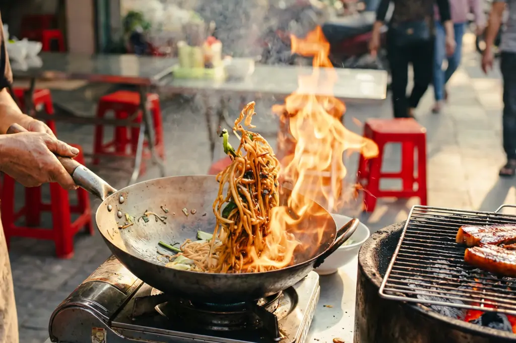 Eye-level medium action shot of phở xào—Vietnamese stir‑fried flat rice noodles—being tossed in a wok over open flames at a street food stall, capturing high heat cooking and wok hei‑style charring.