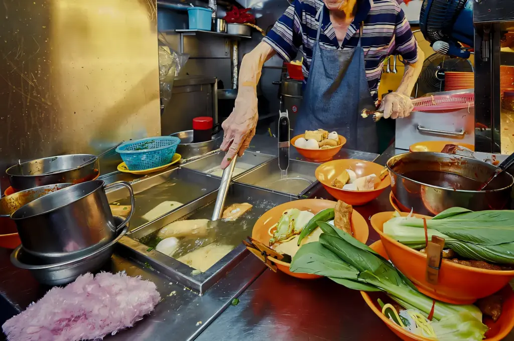 High-angle close-up shot of a hawker ladling hot soup at a stainless steel street food station, surrounded by fresh ingredients and cooking utensils.