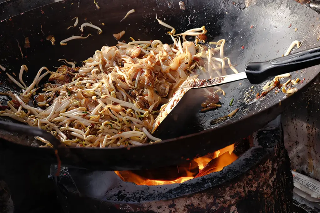Three‑quarter angle close‑up shot of flat rice noodles, bean sprouts, and seasonings being stir‑fried in a hot wok over an open flame while cooking char kway teow.