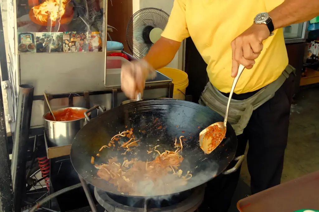 Low-angle action shot of a street food hawker stir-frying char kway teow noodles in a large wok over high heat, with flames, smoke, and ingredients tossing during traditional hawker-style cooking.