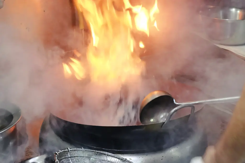 Close-up eye-level shot of a wok blazing over an open flame, with fire and smoke surrounding a metal ladle, illustrating intense heat used in wok hei cooking.