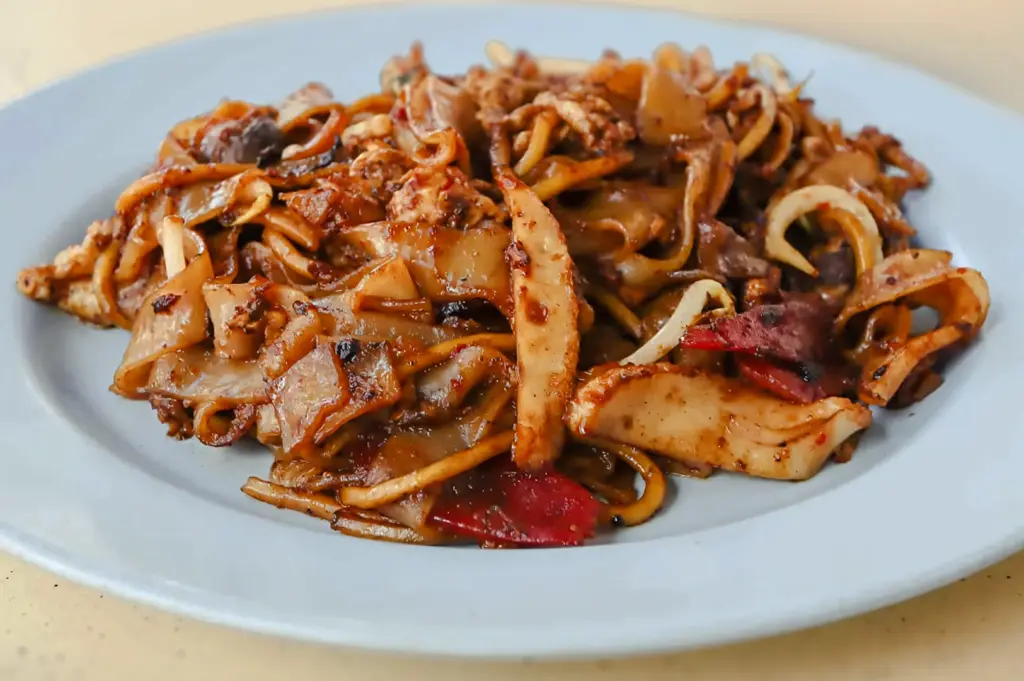 Eye‑level close‑up shot of a plated char kway teow dish with flat rice noodles, cockles, sliced ingredients, and dark soy sauce, served on a light‑colored plate.