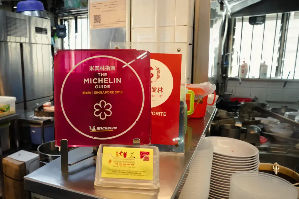 Eye-level close-up shot of a Michelin Guide Singapore award plaque displayed on a hawker stall counter, with kitchen equipment and bowls in the background.